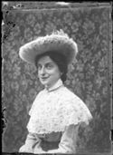 Ritratto di Lina Guerrini sorridente con un cappello di tulle e un abito a quadretti con una mantellina di merletto, davanti a un paravento ricoperto di stoffa con motivo floreale