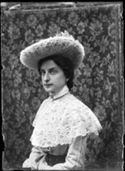 Ritratto di Lina Guerrini con un cappello di tulle e un abito a quadretti con una mantellina di merletto, davanti a un paravento ricoperto di stoffa con motivo floreale
