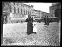 Una donna con una bambina tenuta per mano e due tramway a cavallo in sosta in piazza Vittorio Emanuele II: prospetto del Palazzo Comunale e della fontana del Nettuno in piazza del Nettuno: Bologna