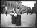 Due donne con un fagotto al braccio fanno conversazione in piazza Vittorio Emanuele II e le persone a passeggio in piazza del Nettuno: prospetto delle vie dell’Indipendenza e Rizzoli: Bologna