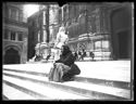 Un’ anziana donna seduta sulla scalinata, una donna di spalle sul sagrato e le persone sedute sullo zoccolo del lato sinistro della basilica di San Petronio: piazza Vittorio Emanuele II: Bologna
