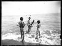 Due bambini di spalle che guardano un bambino con un modellino di barca a vela in legno nella riva: Bellaria