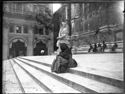 Un’ anziana donna seduta sulla scalinata, una donna di profilo sul sagrato e le persone sedute sullo zoccolo del lato sinistro della basilica di San Petronio: piazza Vittorio Emanuele II: Bologna