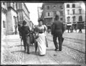 Un anziano uomo, una donna e un uomo di spalle a passeggio in piazza del Nettuno: prospetto delle vie dell’Indipendenza e Rizzoli: Bologna